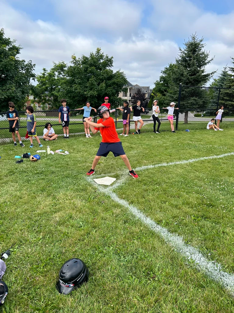 A camper plays baseball with safety helmet, swinging for a pitch, while campers watch on from an outdoor dugout at Rouge Camps Richmond Hill.