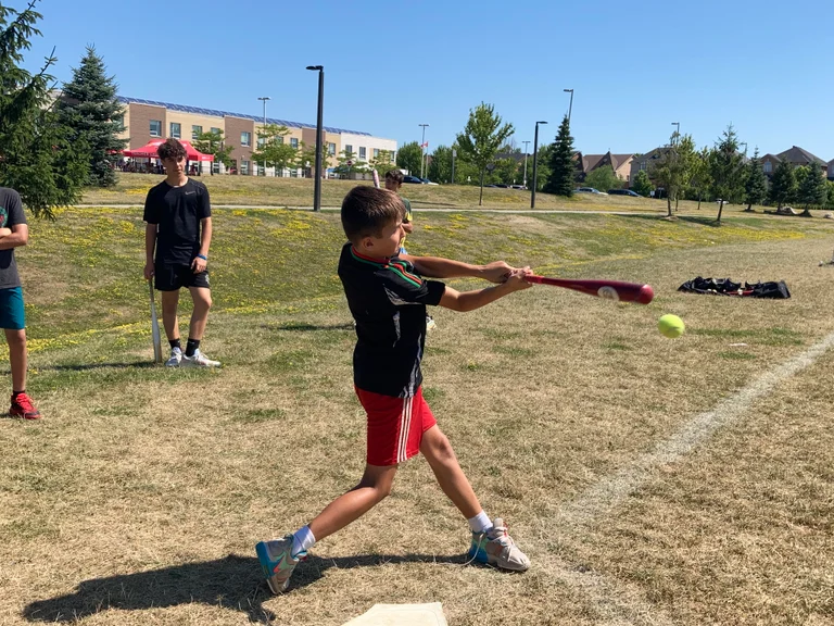 Alt text: A young camper swings a red baseball bat and makes contact with a bright yellow ball on a grassy field. Other campers stand nearby watching, with a school building, trees, and a clear blue sky in the background at Rouge Camps Richmond Hill.