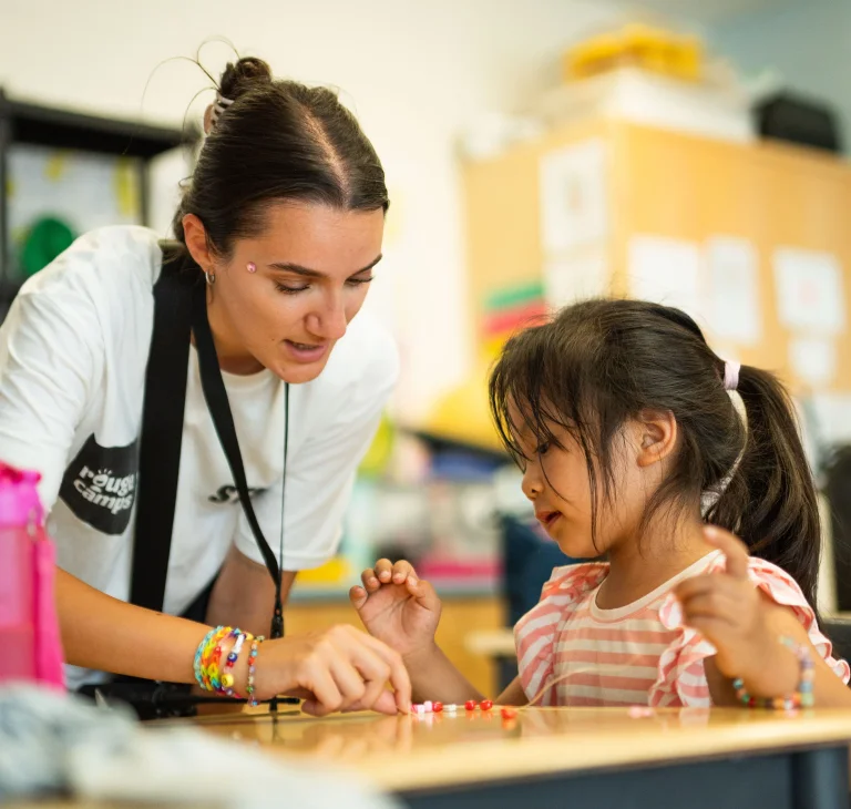 A camp counsellor helps a camper string beads onto a string at Rouge Camps Richmond Hill.