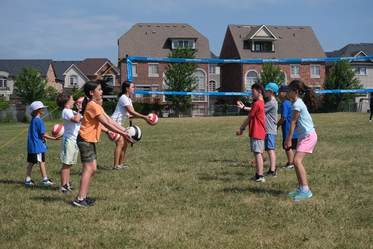 A group of campers stand in two lines on a grassy field playing a volleyball-style passing game. A counsellor in a white shirt demonstrates how to pass a red-and-white ball under a net set up in front of suburban houses at Rouge Camps Richmond Hill.