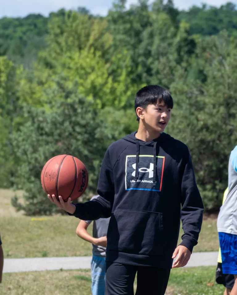 A teen boy in a black hoodie holds a basketball while walking on a paved path during outdoor camp activities, with other campers and trees in the background at Rouge Camps.