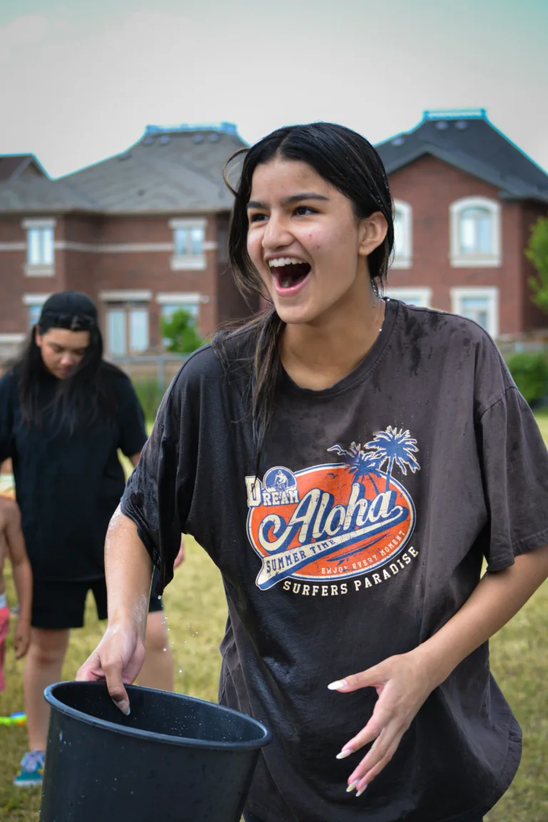 A teenage girl in a wet brown “Aloha” T-shirt laughs while holding a large black bucket during a water activity, with houses and campers in the background at Rouge Camps Richmond Hill.
