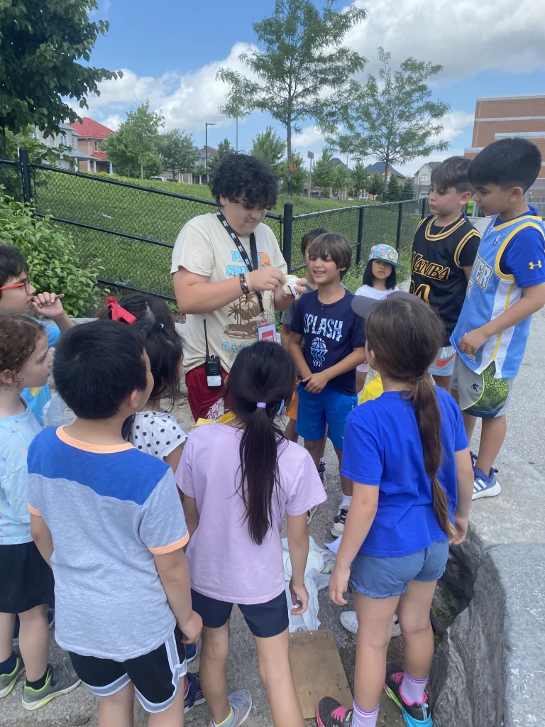 Campers gather around, eagerly looking on as a counsellor explains a science experiment during an educational period at Rouge Camps Richmond Hill.