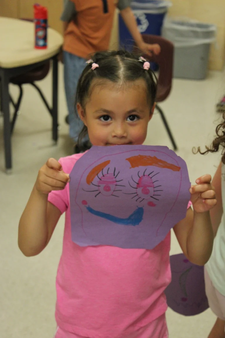 A child smiles while holding up a colorful paper face they created during an art activity at Rouge Camps.