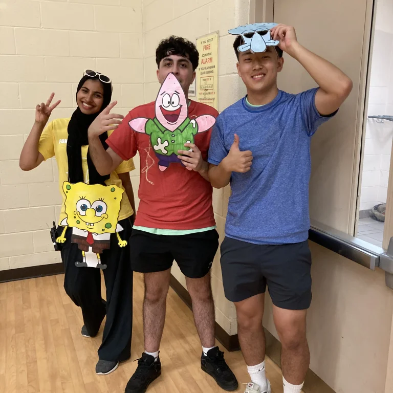 Three camp staff members pose indoors wearing SpongeBob-themed cutouts, smiling and giving peace signs and thumbs up while standing against a gym wall Alt text: Three camp staff members pose indoors wearing SpongeBob-themed cutouts, smiling and giving peace signs and thumbs up while standing against a gym wall at Rouge Camps.