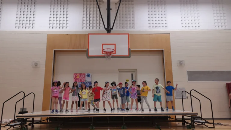 A group of children stand together on a stage in a gym, performing a dance during a camp event at Rouge Camps Woodbridge.