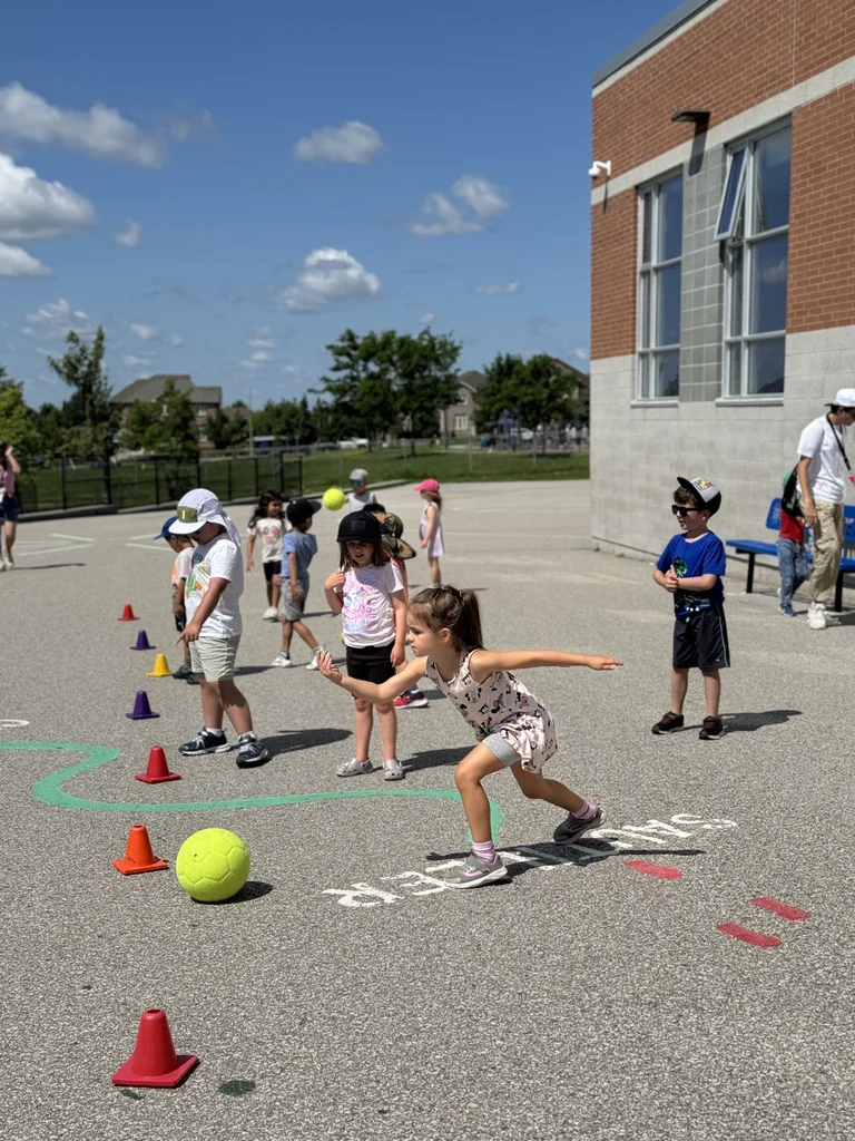 A child reaches toward a green ball during an outdoor game, with other children and cones set up on a sunny schoolyard at Rouge Camps Richmond Hill.