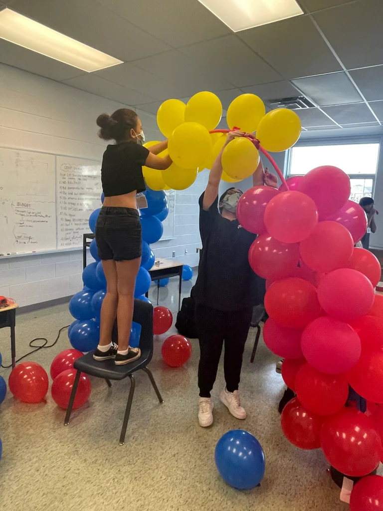 Two staff members assemble a colorful balloon arch indoors. One stands on a chair attaching yellow balloons while the other holds the red and yellow arch frame. Red and blue balloons are scattered on the classroom floor at at Rouge Camps Richmond Hill.