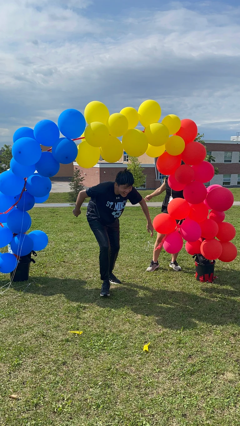 A teenage boy runs forward under a large balloon arch made of blue, yellow, and red balloons set up on a grassy field. A school building and cloudy sky are visible in the background at Rouge Camps Richmond Hill.