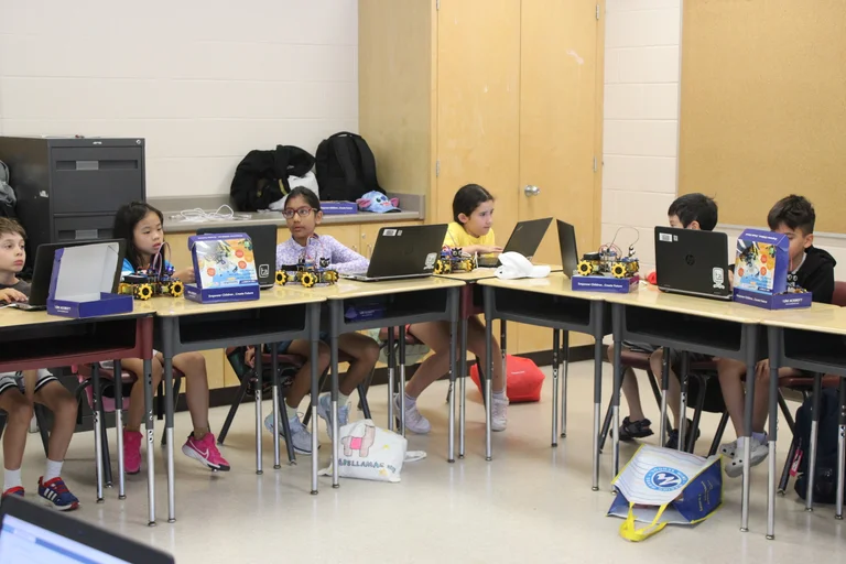 A group of students sit at desks using laptops and robotics kits in a classroom at Rouge Camps.
