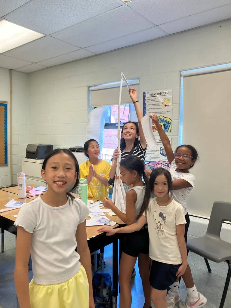A group of children smile and work together on a tall paper structure during an indoor classroom activity at Rouge Camps.