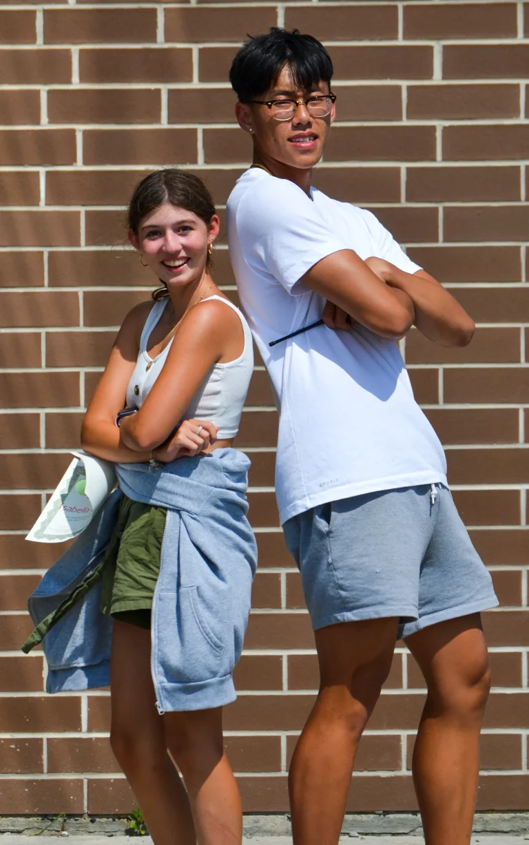 One teen camper and her counsellor stand back-to-back against a brick wall outdoors, smiling with arms crossed in a playful, confident pose at Rouge Camps Richmond Hill.