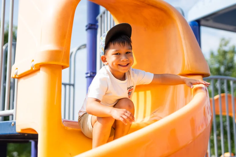 A young camper smiles while sitting at the top of a bright orange playground slide, reaching forward and ready to slide down on a sunny day at Rouge Camps.