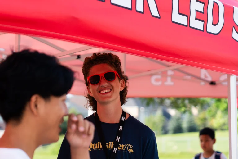 A teen counsellor wearing red sunglasses smiles under a red Rouge Camps tent while talking with another person in the foreground at Rouge Camps