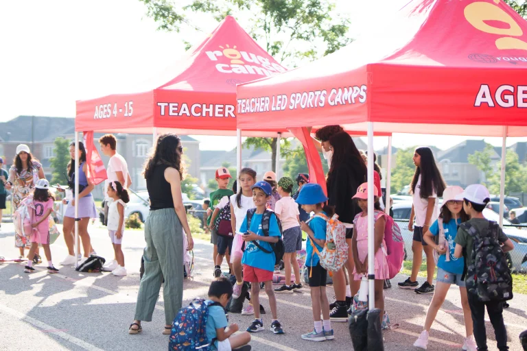 Campers with backpacks gather under red Rouge Camps tents labeled “Teacher Led Sports Camps” and “Ages 4–15” during morning drop-off, as staff greet families on a sunny day at Rouge Camps Richmond Hill.