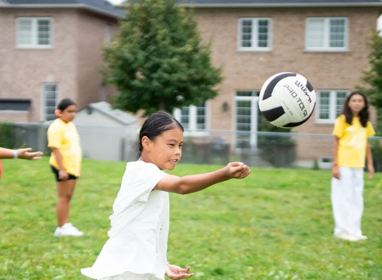 A camper in a white outfit bumps a volleyball on a grassy field while other campers and a counsellor watch nearby, with houses in the background at Rouge Camps Richmond Hill..