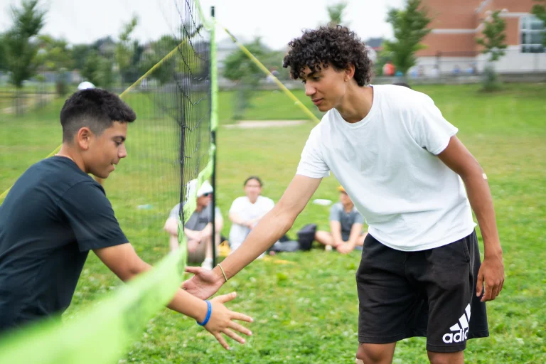 Two campers reach across a volleyball net to high-five during an outdoor camp activity on a grassy field, with other campers sitting and watching in the background at Rouge Camps.