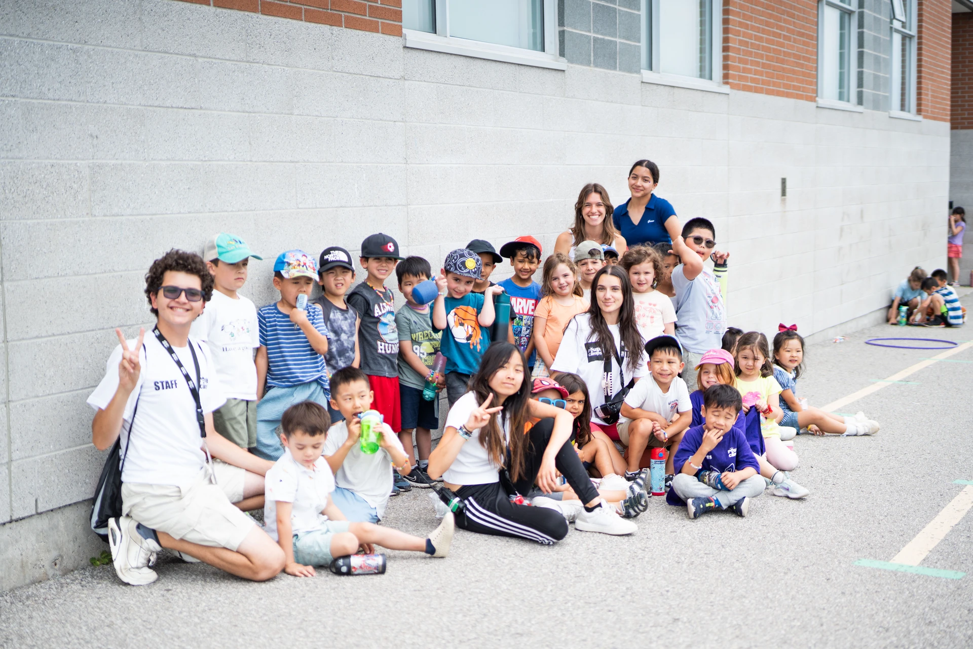 A group of Little Movers campers poses outdoors, beaming with smiles and in front of their caring staff members at Rouge Camps Richmond Hill.