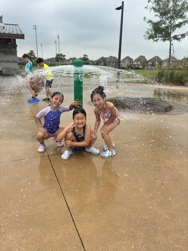 A group of three female campers smile ear to ear while hiding under a mushroom sprinkler during water play of a hot summer day at Rouge Camps Woodbridge.