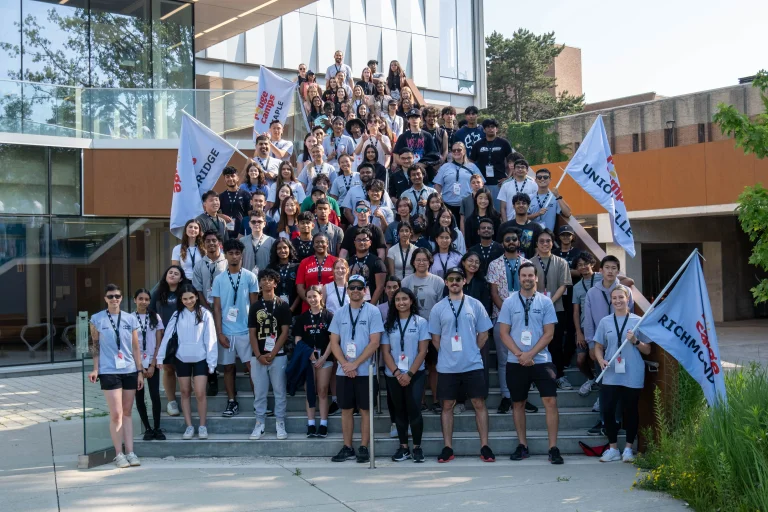 A large group of Rouge Camps staff and leaders pose together on outdoor steps in front of a modern building, holding location flags and smiling during a team gathering.